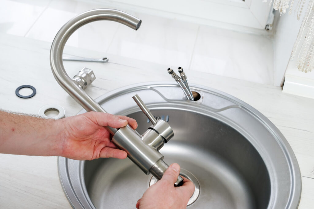 A person installs a modern metal kitchen faucet over a round sink, with plumbing parts and tools visible on the countertop.