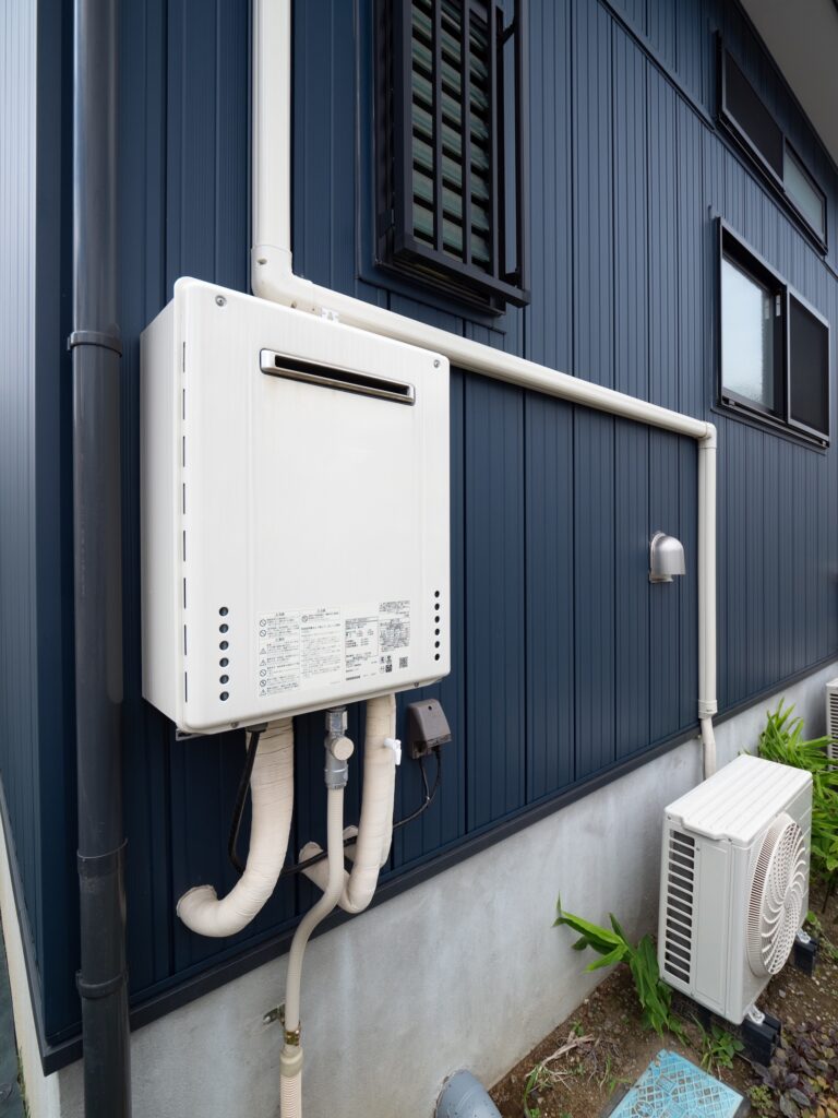 A white outdoor tankless water heater is mounted on a blue exterior wall near an air conditioning unit and several pipes.