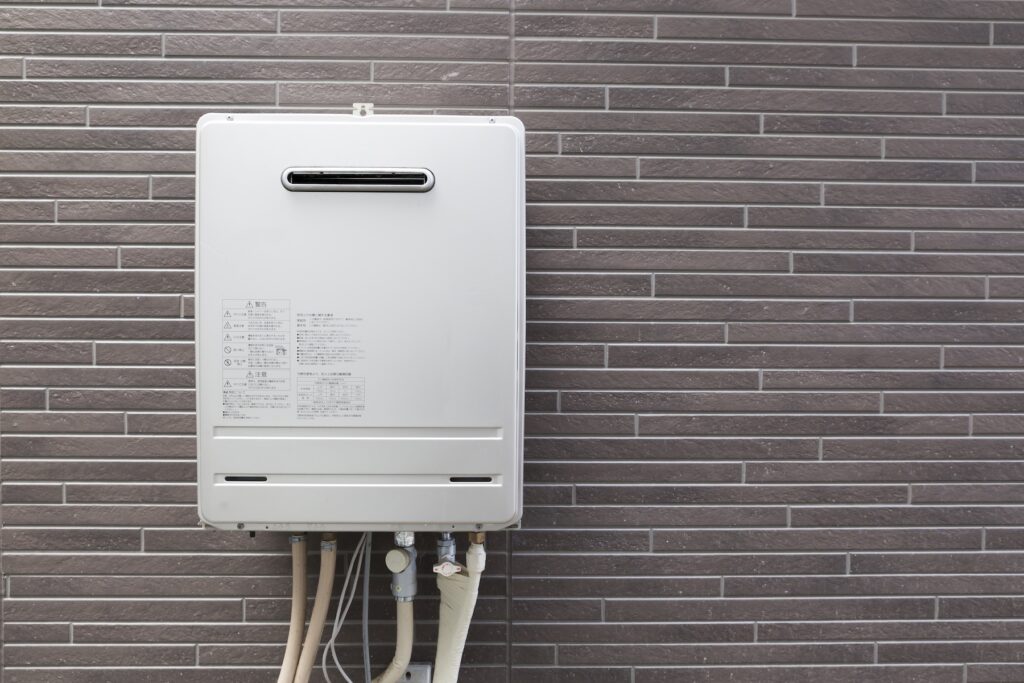 A white tankless water heater mounted on a gray tiled wall with several pipes connected at the bottom.